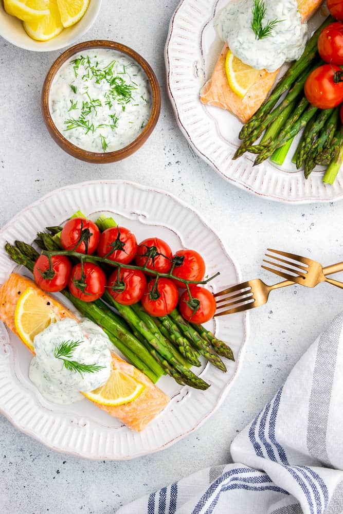 Baked Salmon topped with dill sauce, asparagus and cherry tomatoes on a vine, served on a white plate.