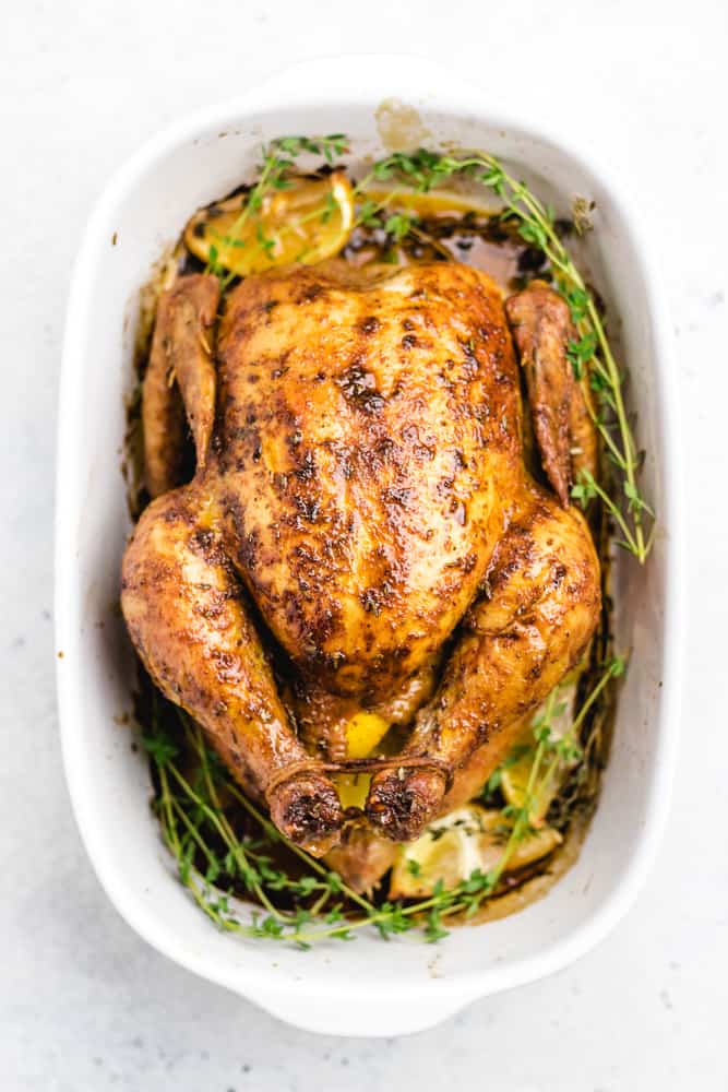 A cooked whole roasted chicken placed in a white baking pan with thyme and lemin wedges.