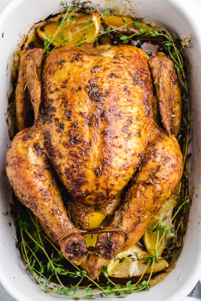 A cooked whole roasted chicken placed in a white baking pan with thyme and lemin wedges.