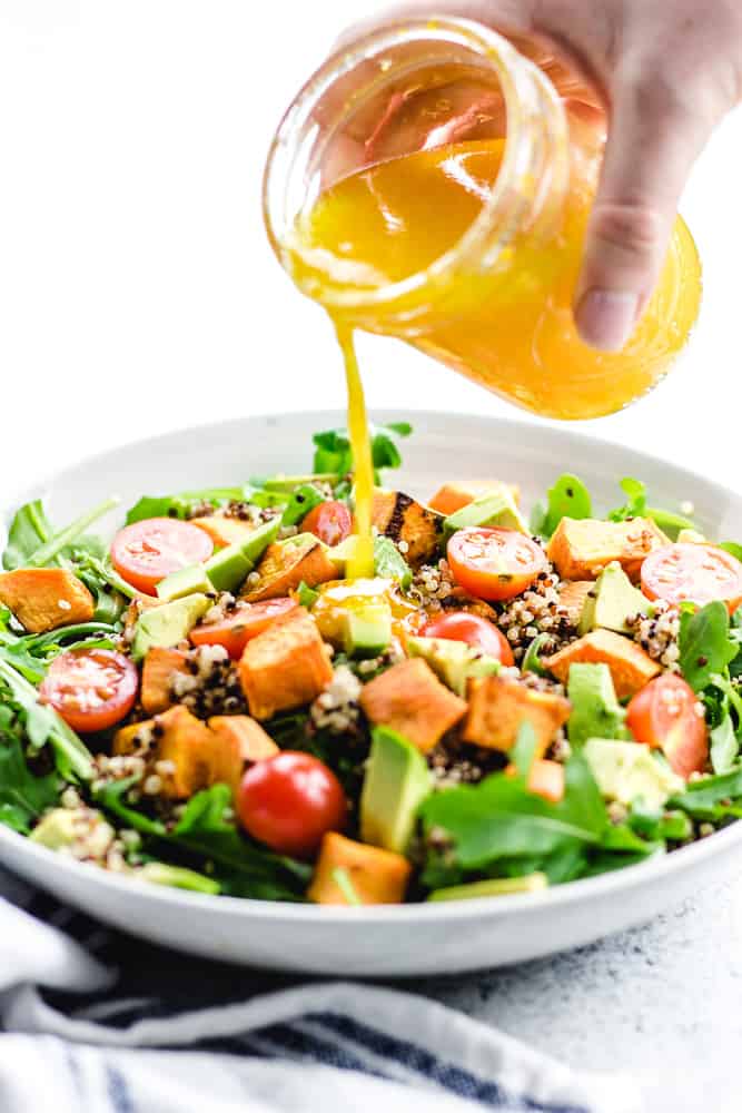 Pouring orange dressing over salad with quinoa, arugula, sweet potatoes, avocado, and tomatoes.