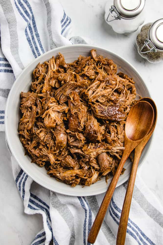 A bowl with pulled pork and two woden spoons on a white table.