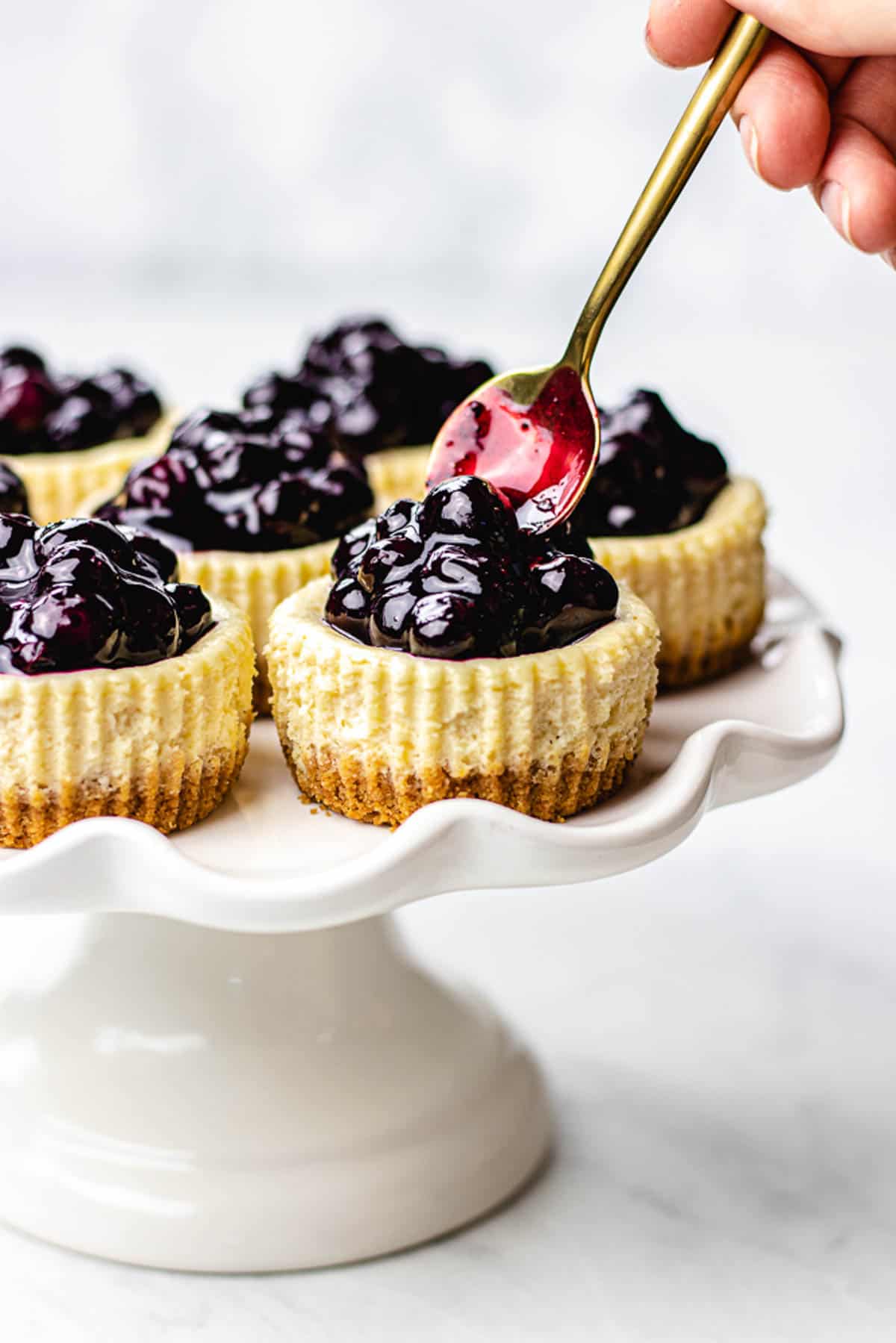 seven mini cheesecakes with blueberry sauce on a white cake plate.
