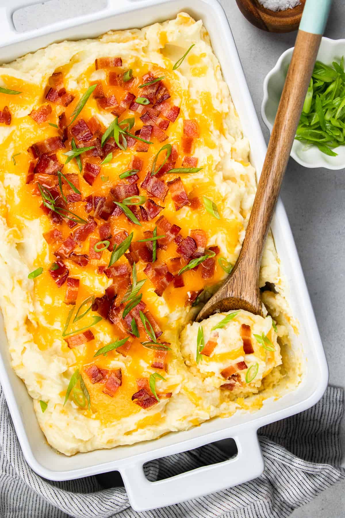 Mashed potatoes topped with cheese and bacon in a white casserole with a wooden spoon.