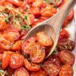 Roasted halved red cherry tomatoes in a white baking pan with a wooden spoon.