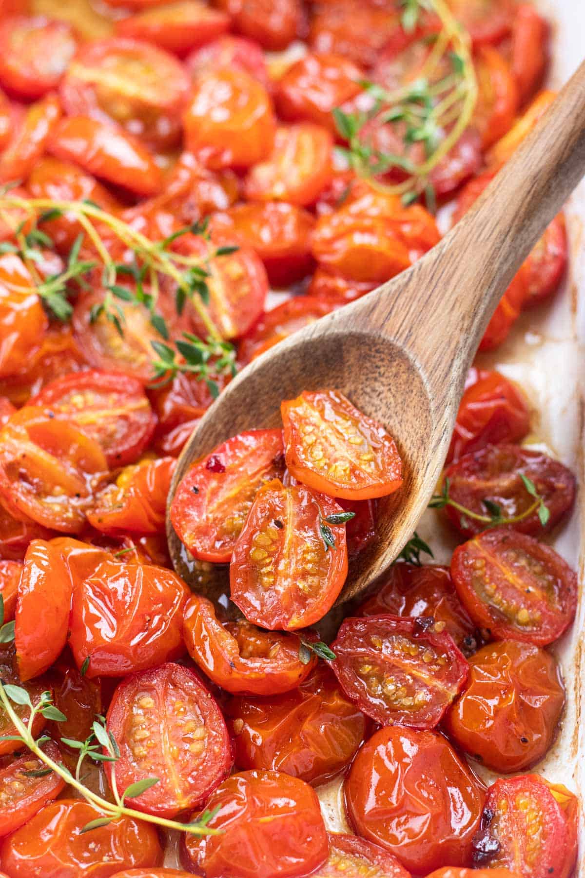 Roasted halved red cherry tomatoes in a white baking pan with a wooden spoon.