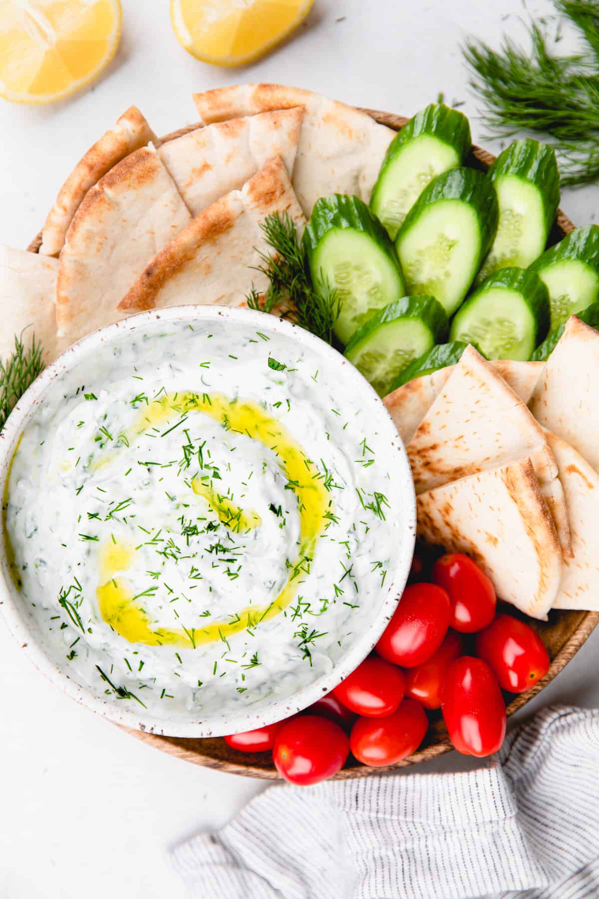 Tzatziki sauce, topped with olive oil and chopped dill, in a white bowl and vegetables and pita bread around it.