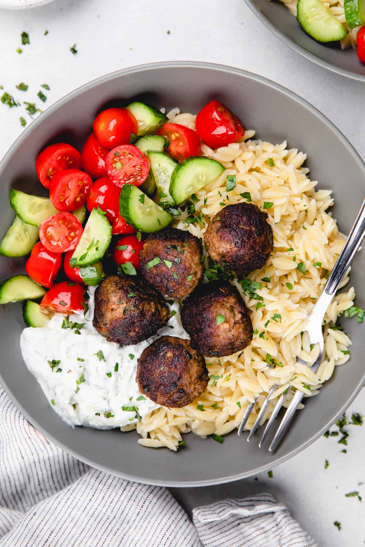 Greek meatballs with orzo, tzatziki sauce, and cherry cucumber salad in a grey bowl with a fork.