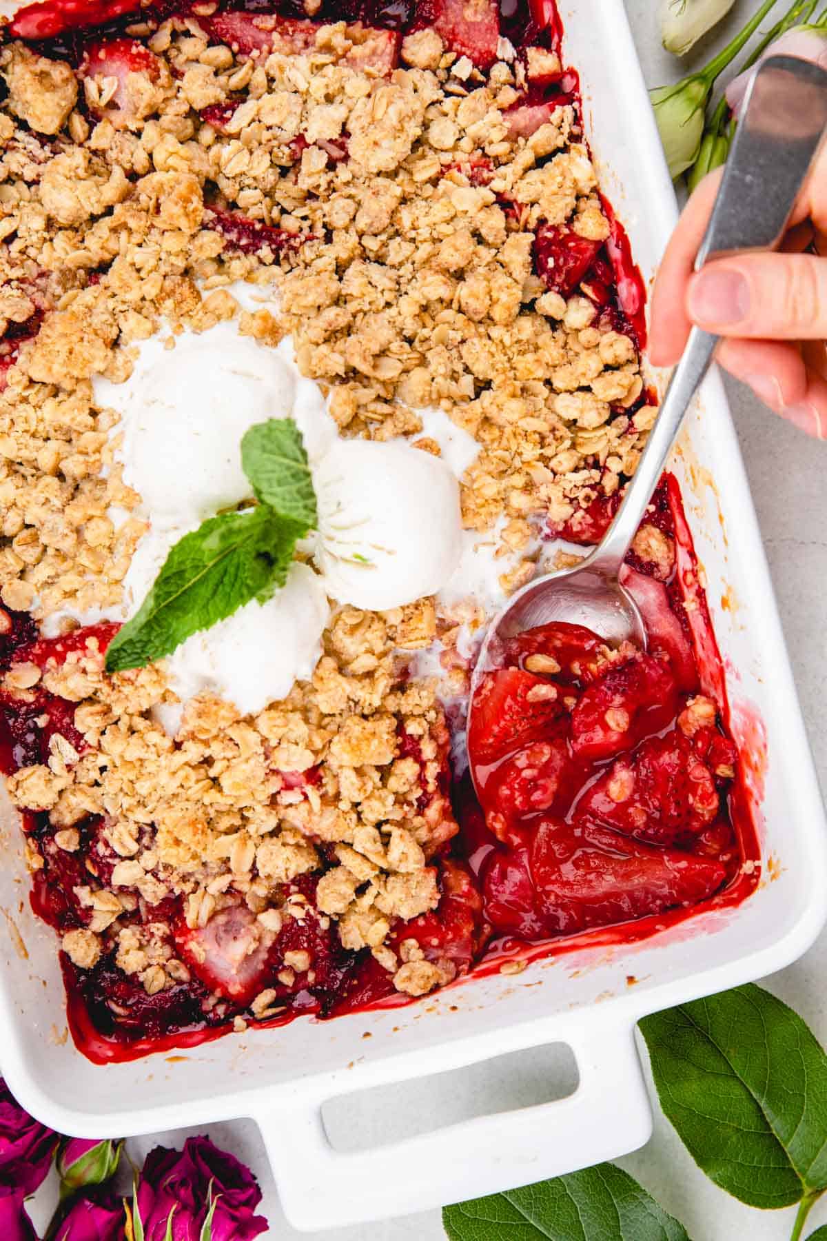 Scooping out strawberry crumble with a spoon, topped with ice cream and mint leaves.