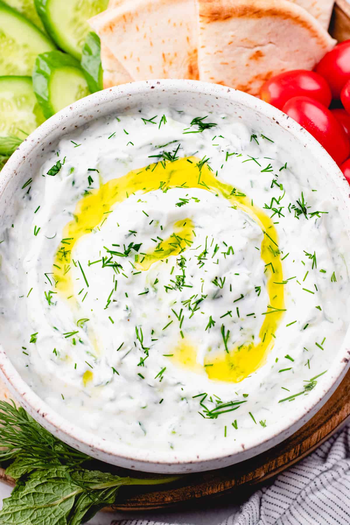Tzatziki sauce, topped with olive oil and chopped dill, in a white bowl and vegetables and pita bread around it.
