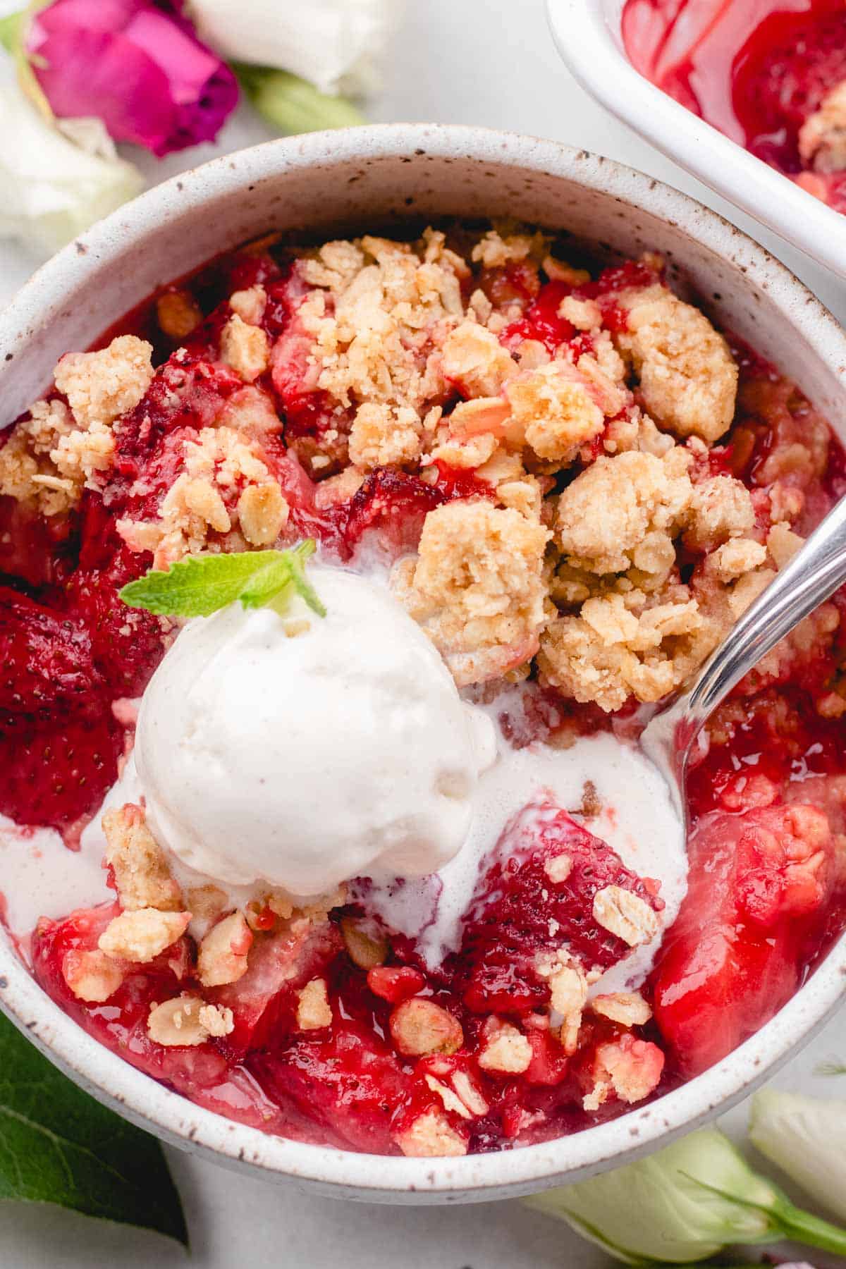 Scooping out strawberry crumble with a spoon, topped with ice cream and mint leaves.