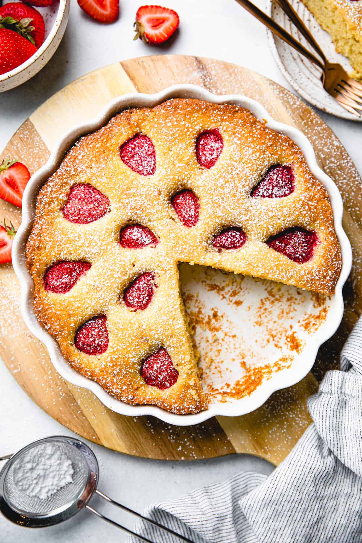 A vanilla coffee cake, topped with strawberries, in a white round baking pan on a wooden board.
