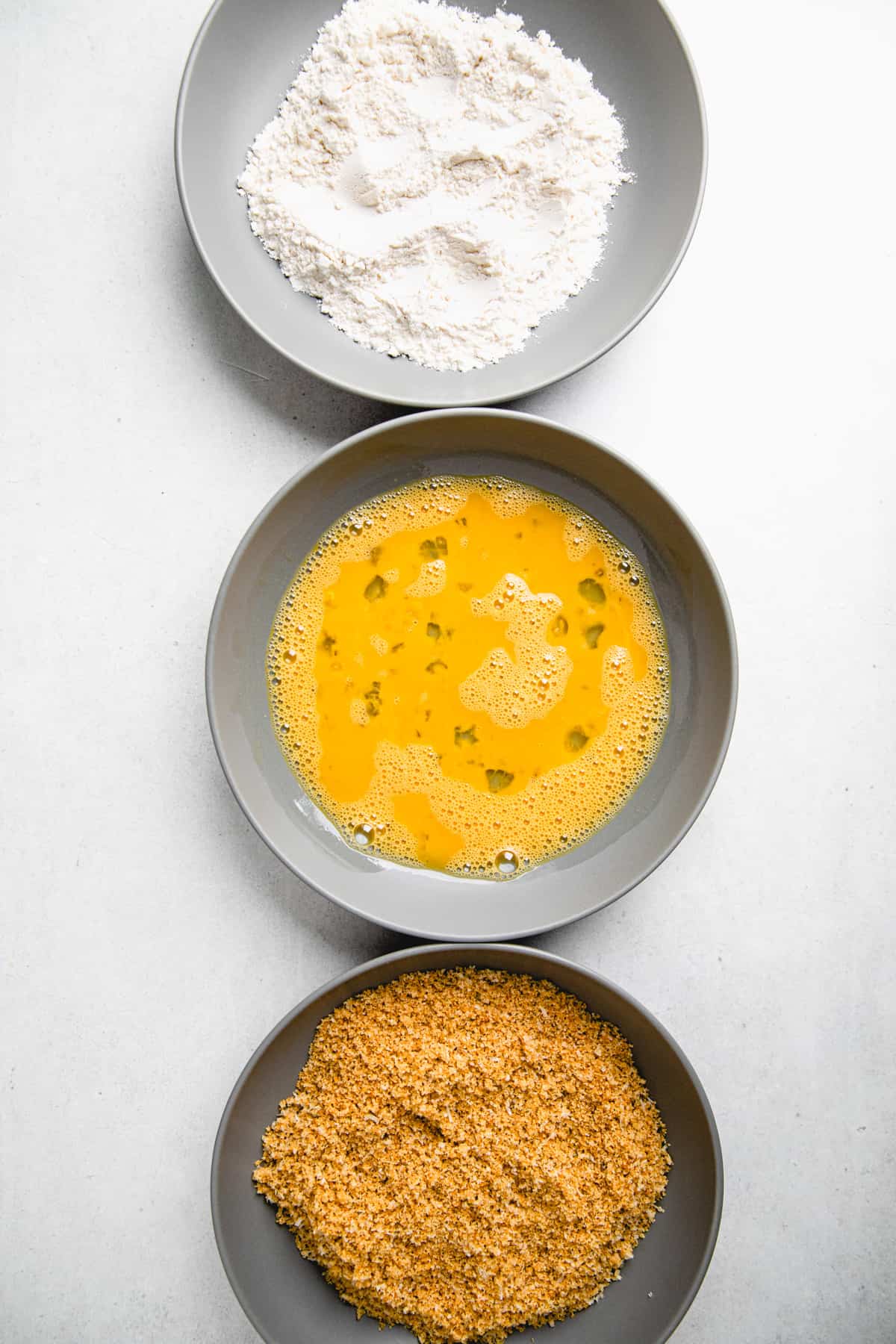 Three grey bowl, filled with flour, beaten eggs, and breadcrumbs.