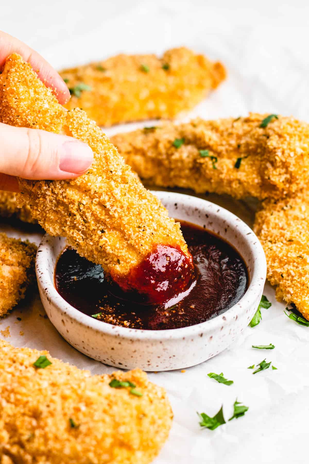 Baked chicken tenders on parchment paper with BBQ sauce in a white bowl.