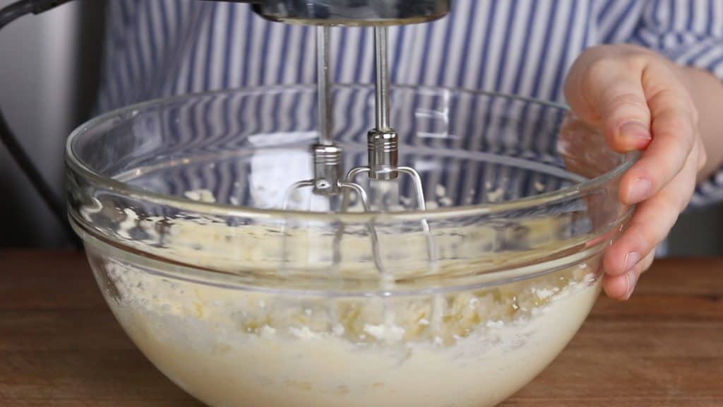 A process photo of mixing ingredients with a hand mixer in a glass bowl.