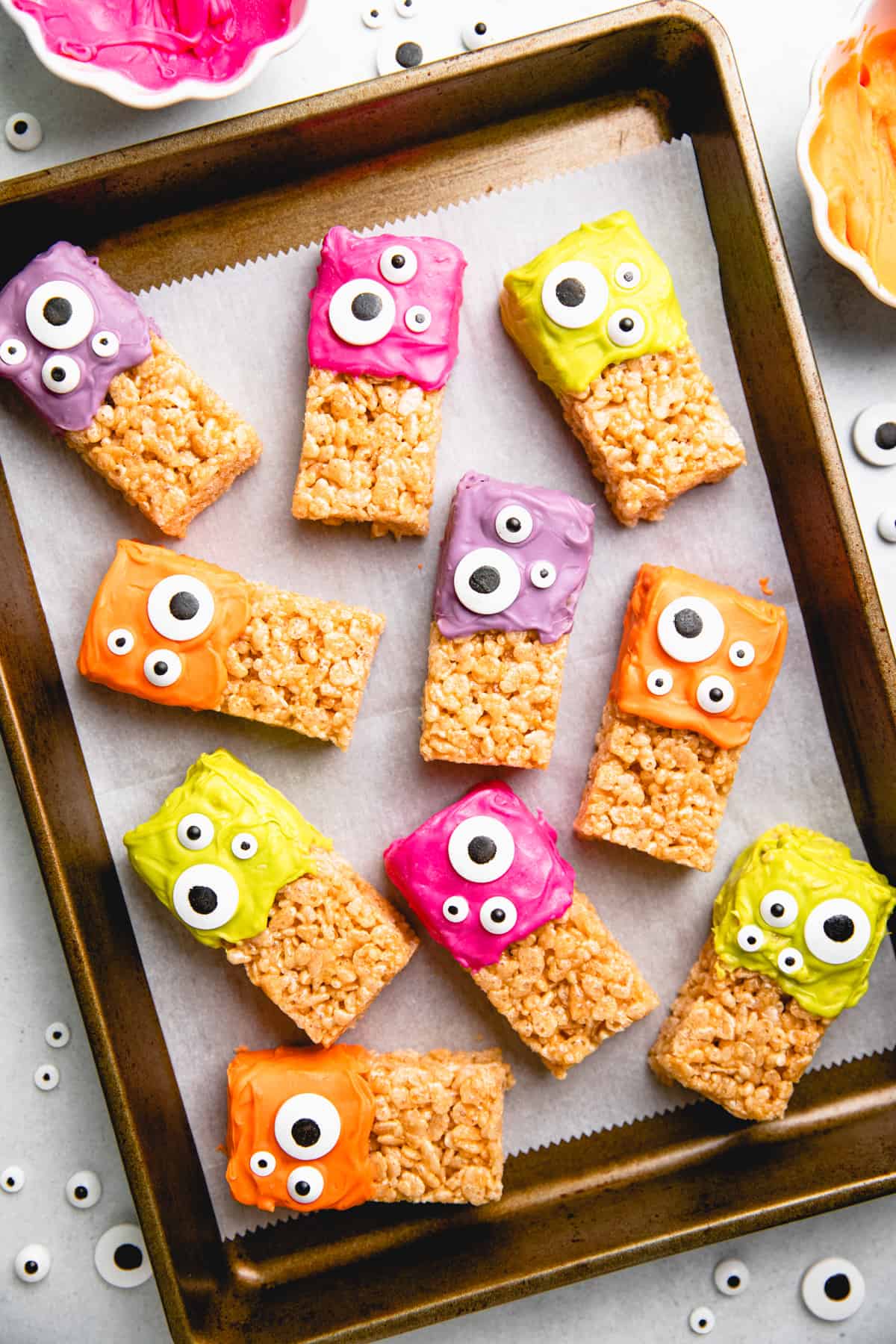 Halloween Rice Krispie Treats, topped with colored icing and candy eyes on baking sheet with parchment paper.