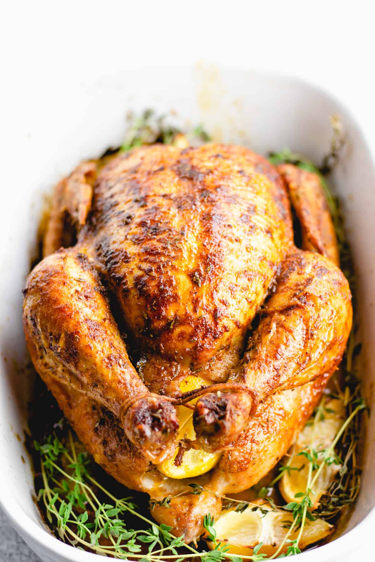 A cooked whole roasted chicken placed in a white baking pan with thyme and lemon wedges.