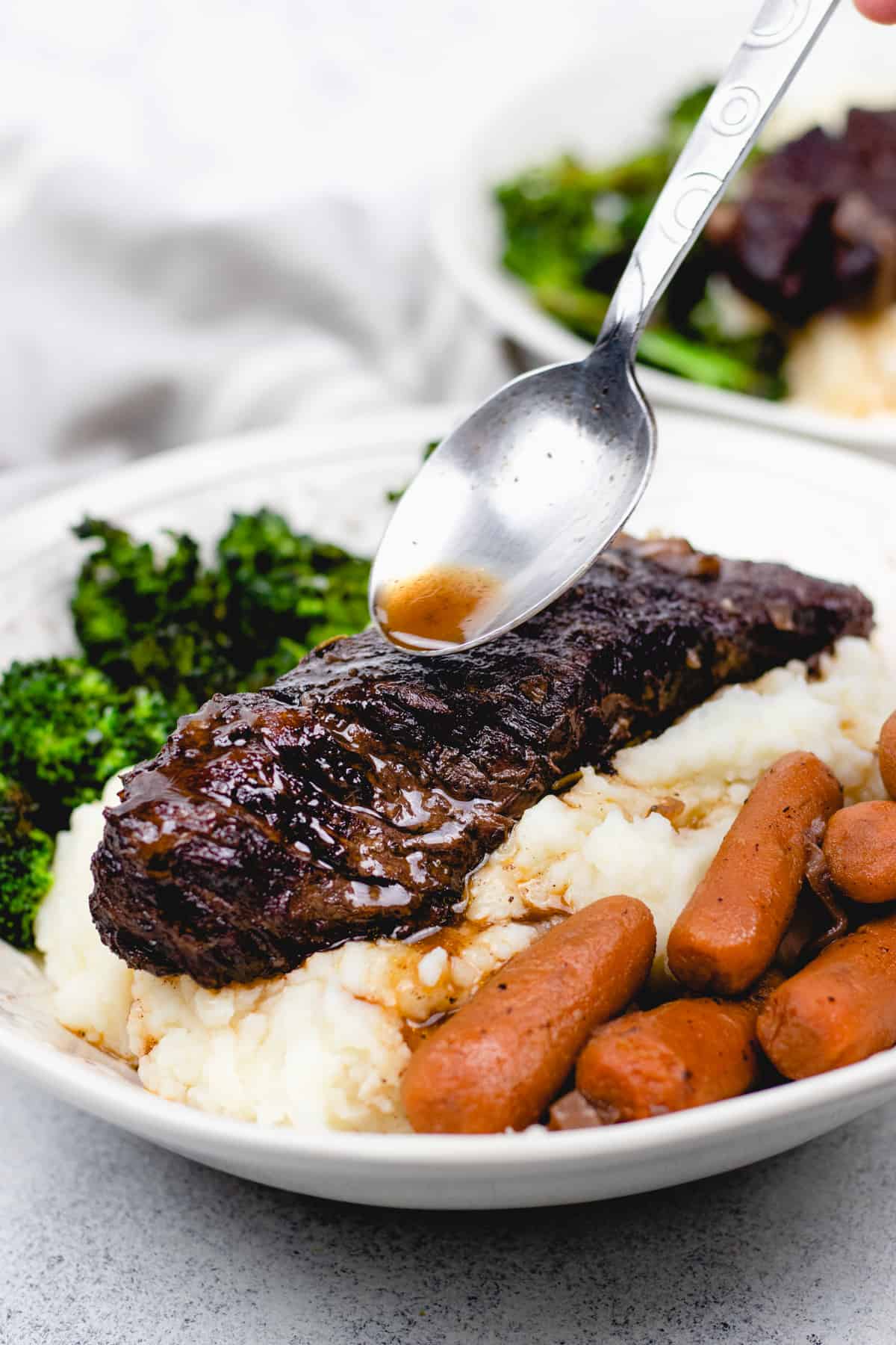 Pouring gravy over Boneless short rib, mashed potatoes, broccoli, and carrots in a bowl.