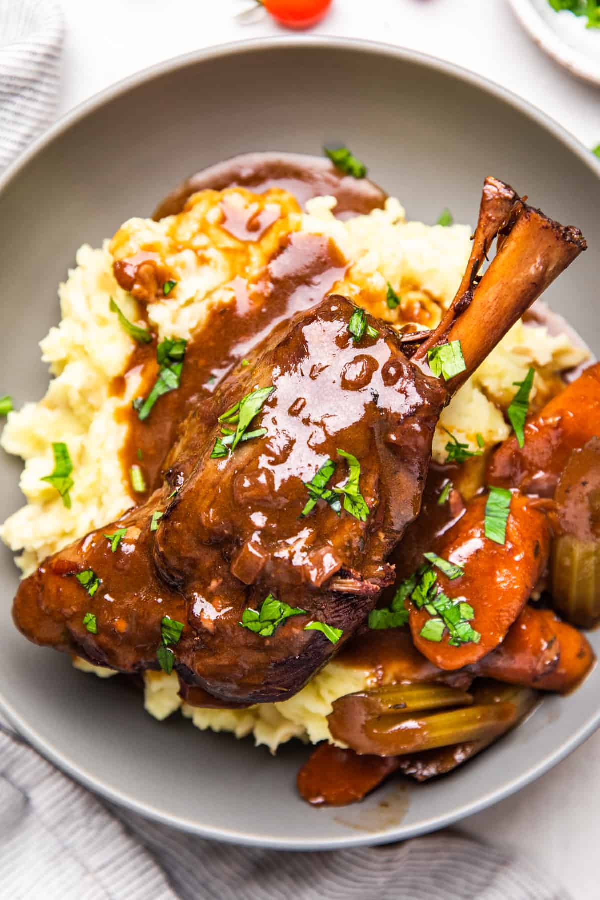 Lamb shank with gravy, vegetables, amd mashed potatoes in a grey bowl.