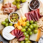 A serving board, topped with cured meats, cheese, crackers, grapes, nuts, and jam.