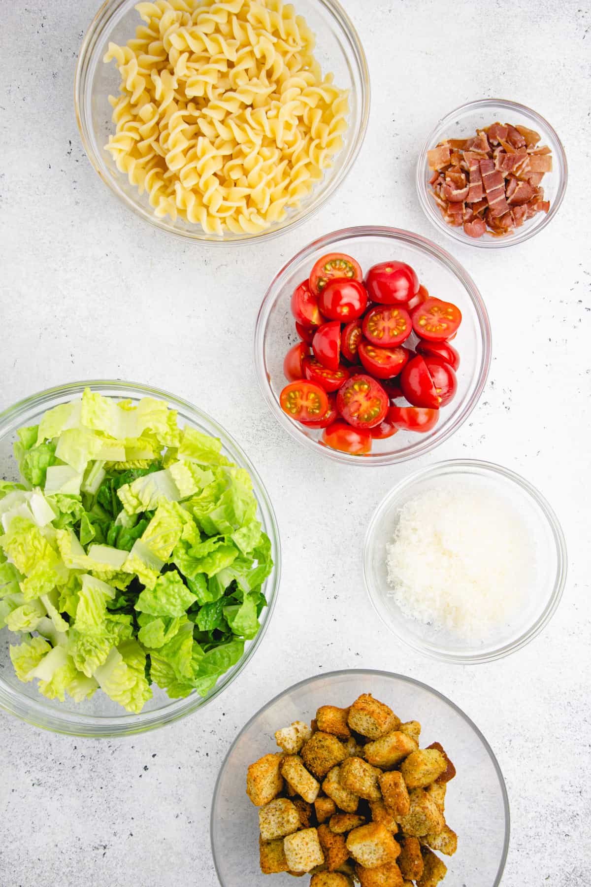 Chopped lettuce, cherry tomatoes, bacon, pasta, croutons, and Parmesan cheese in separate glass mixing bowls.