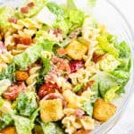 Salad with lettuce, pasta, cherry tomatoes, bacon, and croutons in a glass mixing bowl.