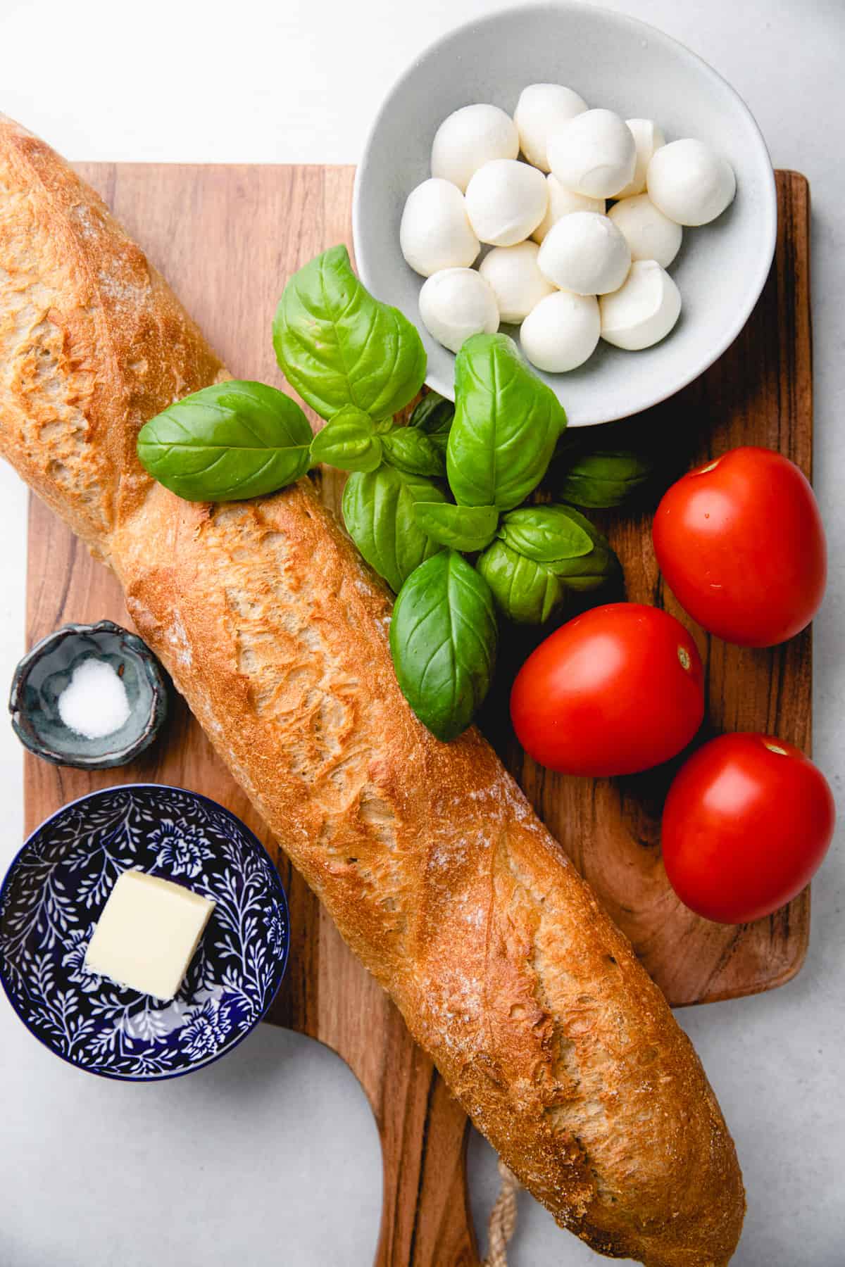 French loaf, Roma tomatoes, mozzarella cheese, and basil on a cutting board.