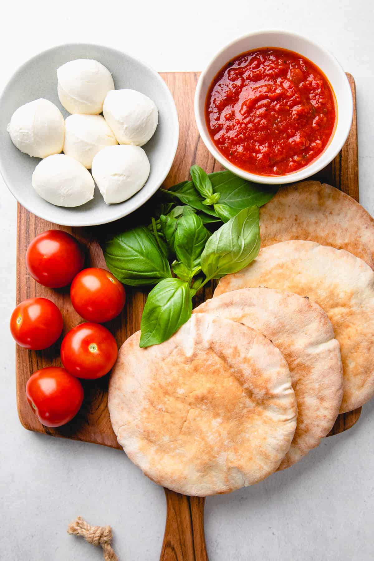 Pita bread, cherry tomatoes, basil, pizza sauce, and mozzarella cheese on a cutting board.