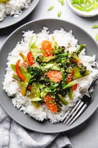 Sauteed broccoli, carrots, snow peas, and red bell pepper in teriyaki sauce over white rice in a bowl.