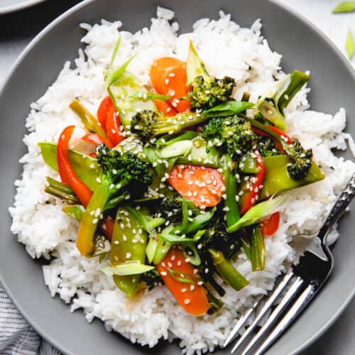Sauteed broccoli, carrots, snow peas, and red bell pepper in teriyaki sauce over white rice in a bowl.