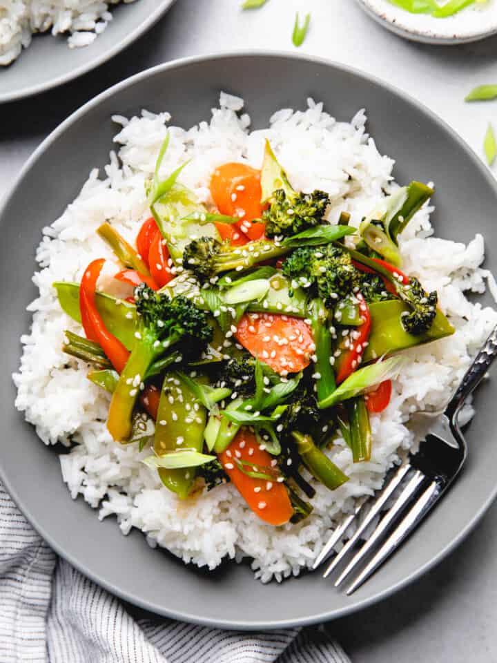 Sauteed broccoli, carrots, snow peas, and red bell pepper in teriyaki sauce over white rice in a bowl.