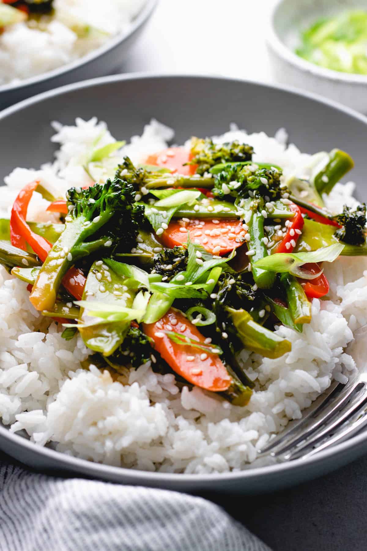 Sauteed broccoli, carrots, snow peas, and red bell pepper in teriyaki sauce over white rice in a bowl.