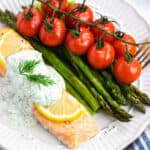 A piece of baked salmon, topped with lemon slices and creamy dill sauce, with asparagus and tomatoes on the side.