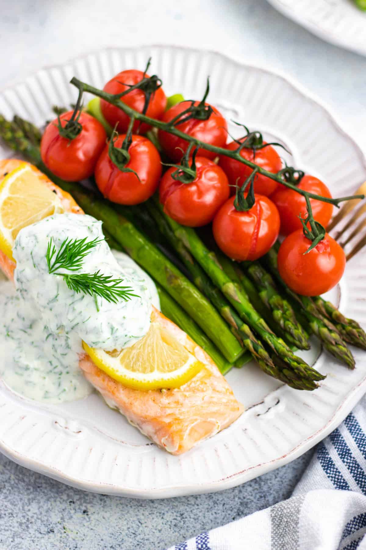 A piece of baked salmon, topped with lemon slices and creamy dill sauce, with asparagus and tomatoes on the side.