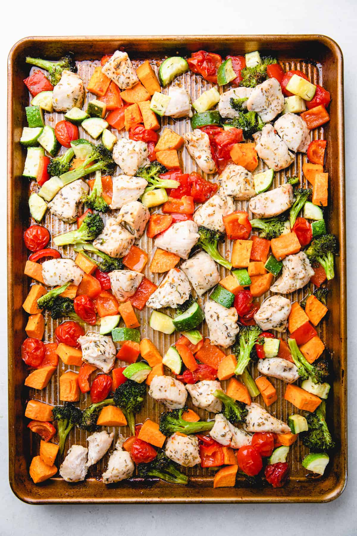 Baked diced chicken breast, sweet potatoes, broccoli, zucchini, bell pepper, and cherry tomatoes on a baking sheet.