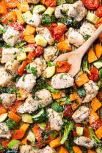 Baked diced chicken breast, sweet potatoes, broccoli, zucchini, bell pepper, and cherry tomatoes on a baking sheet topped with grated Parmesan cheese and herbs.