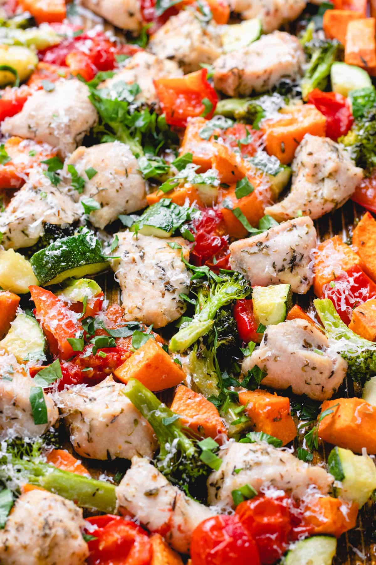 Baked diced chicken breast, sweet potatoes, broccoli, zucchini, bell pepper, and cherry tomatoes on a baking sheet topped with grated Parmesan cheese and herbs.