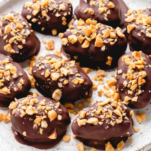 Dates, filled with peanut butter, covered with chocolate, and topped with chopped peanuts, on a white plate.