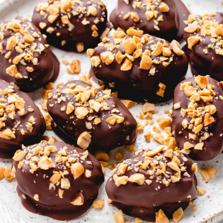 Dates, filled with peanut butter, covered with chocolate, and topped with chopped peanuts, on a white plate.