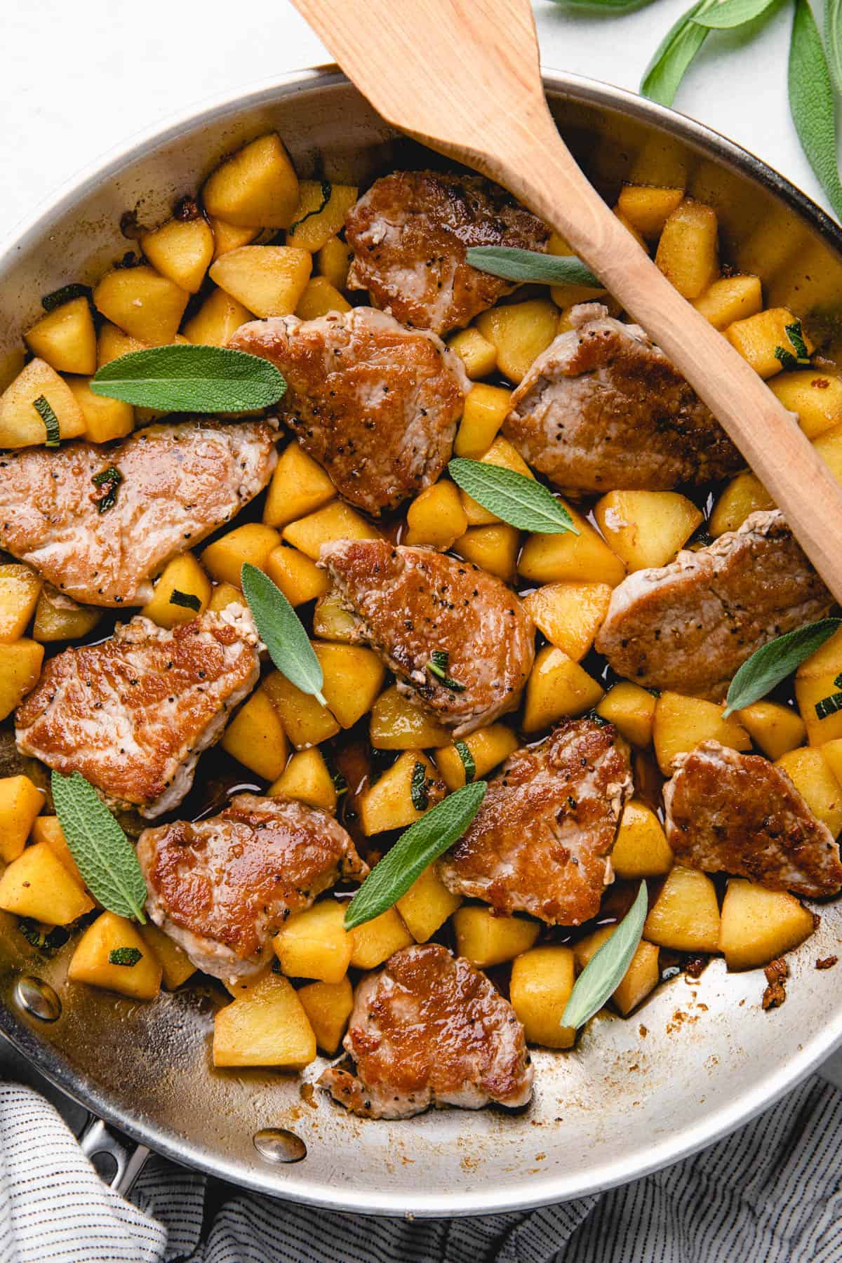 Pork medallions with caramelized apples in a skillet, topped with fresh sage leaves.