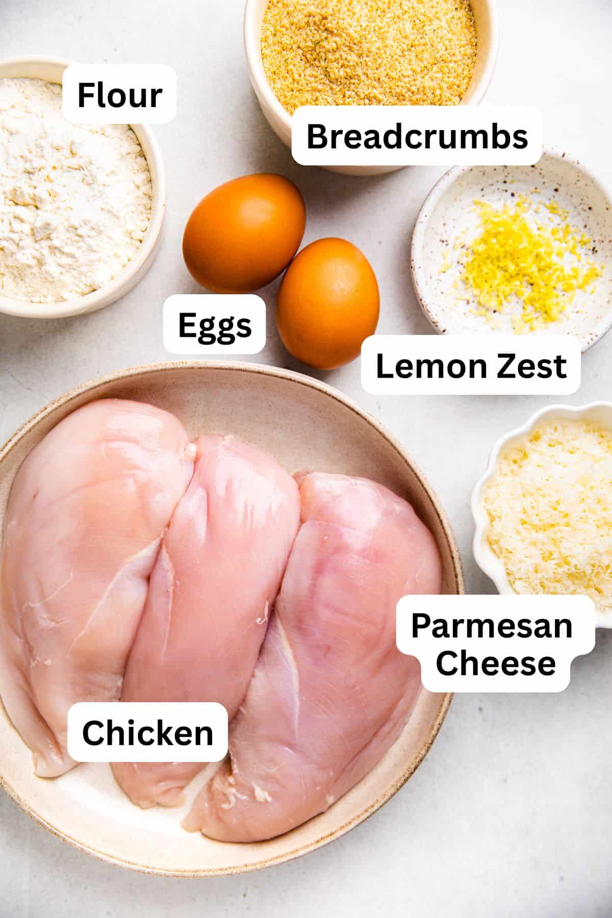 Raw chicken breasts, flour, breadcrumbs, lemon zest, Parmesan cheese, and eggs.